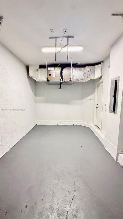 Enclosed storage space featuring painted concrete flooring, wall-mounted electrical panel, ceiling-mounted fluorescent lighting, and overhead HVAC system