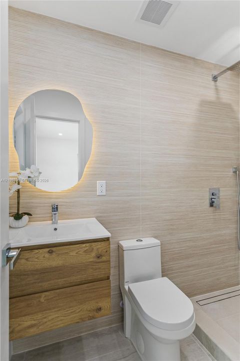 Floating wood-finish vanity with an integrated sink, backlit organic-shaped mirror, wall-mounted shower fixture, and textured stone-finish wall tile
