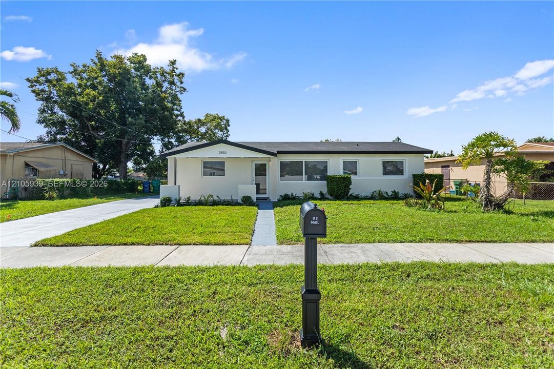 Single-story exterior featuring a light-colored stucco facade, a low-pitched roofline, a paved driveway, and an expansive front lawn