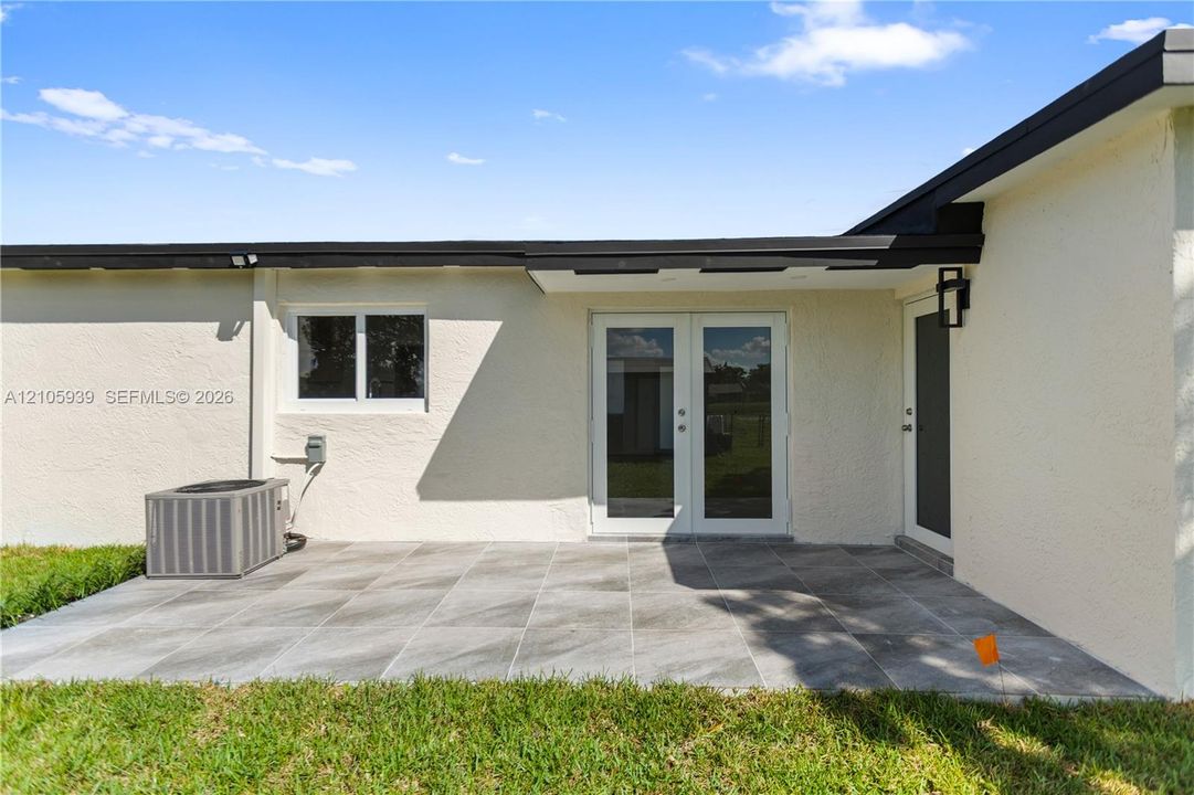 Rear patio featuring large-format gray tile flooring, double-glass entry doors, an attached exterior light fixture, and a stucco finish