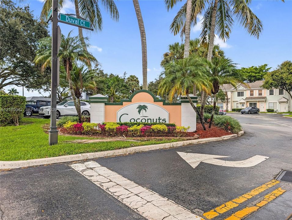 Community entrance sign featuring masonry pillars, integrated landscaping, mature palm trees, and asphalt roadway