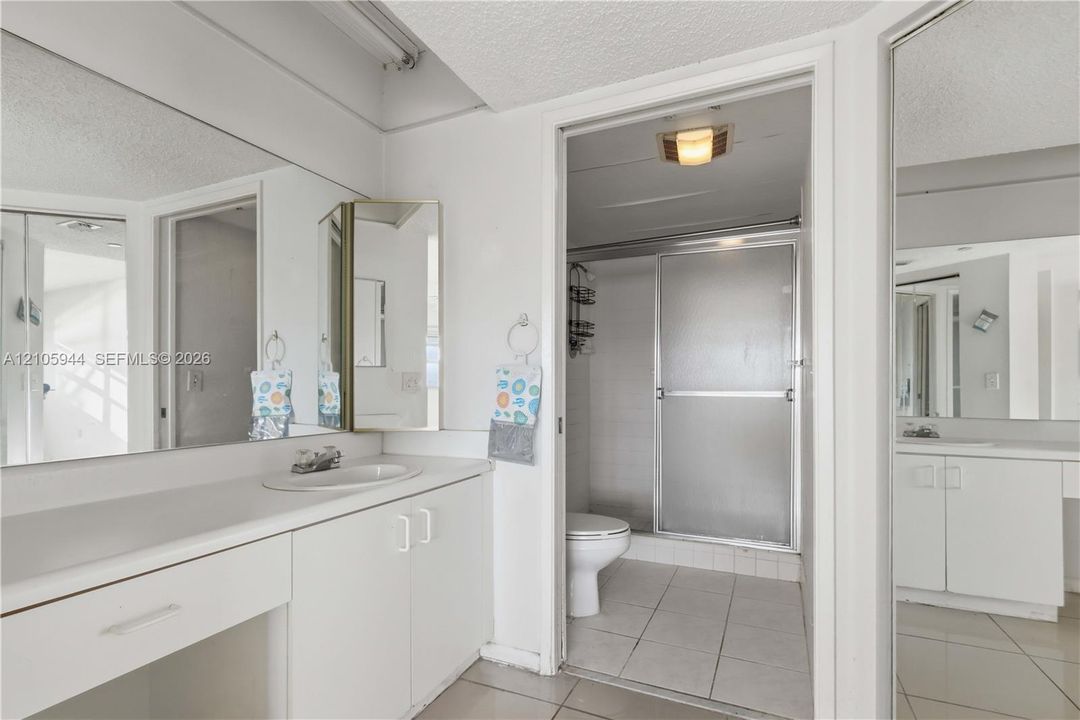 Bathroom featuring a full-width vanity with integrated basin, expansive wall-mounted mirror, white cabinetry, and a separate water closet area with a walk-in shower enclosure and tile flooring