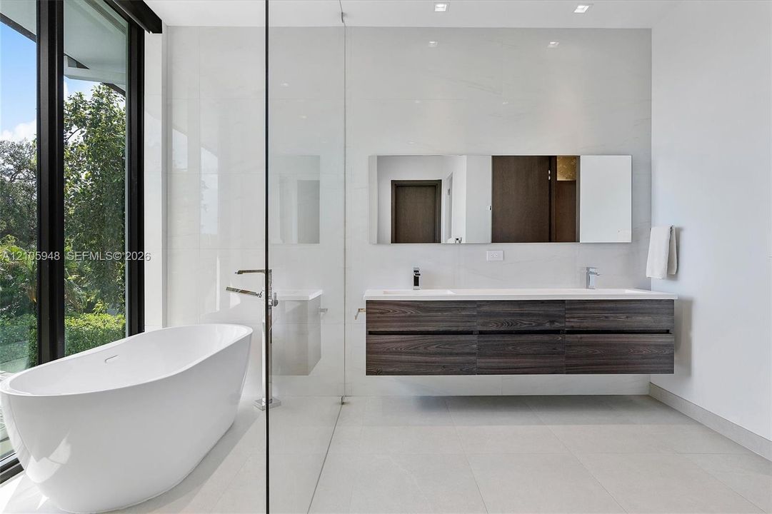 Freestanding oval soaking tub, wall-mounted dark wood-finish dual vanity with integrated sinks, large-format tile flooring, floor-to-ceiling glass-enclosed shower, and expansive window wall