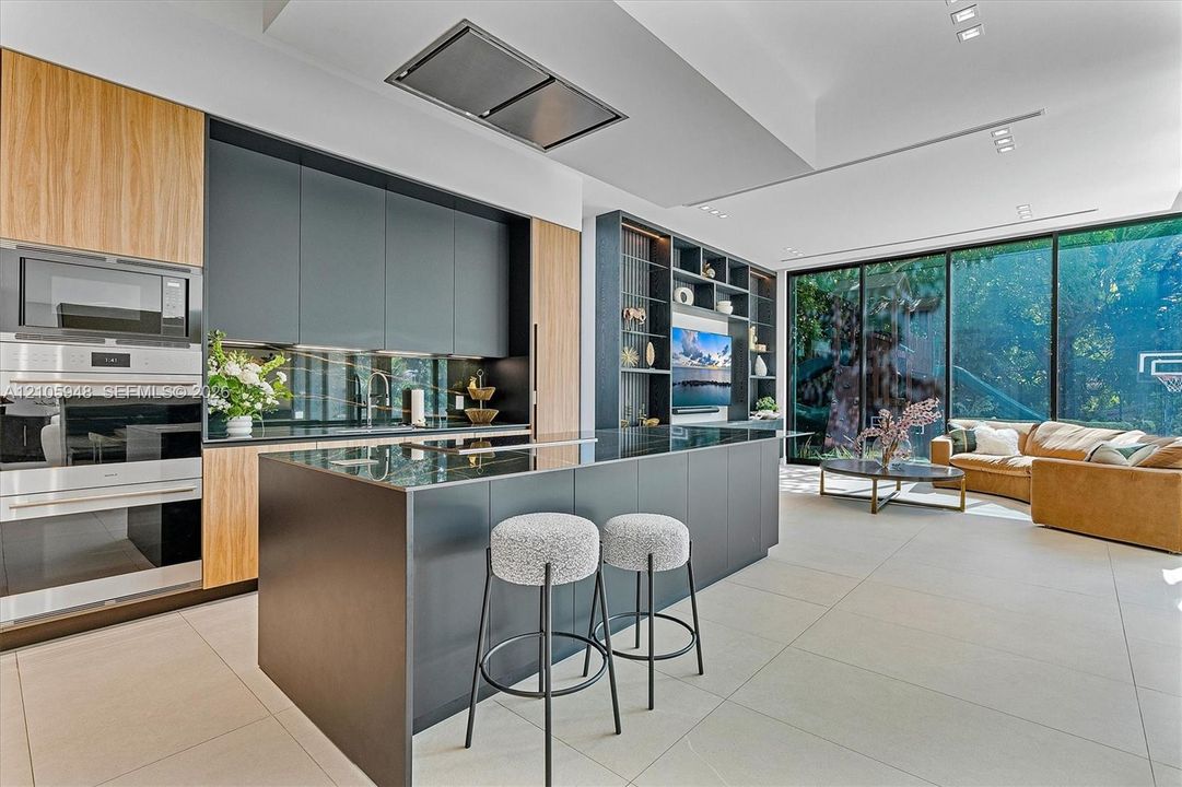 Contemporary kitchen featuring flat-panel cabinetry, a central island with a waterfall edge, stainless steel built-in double wall ovens, ceiling-integrated recessed lighting, and floor-to-ceiling glass wall panels
