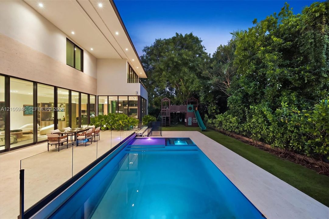 Rear exterior featuring floor-to-ceiling glass wall panels, stone-finish patio, rectangular swimming pool with integrated spa, glass railing, and manicured landscaping