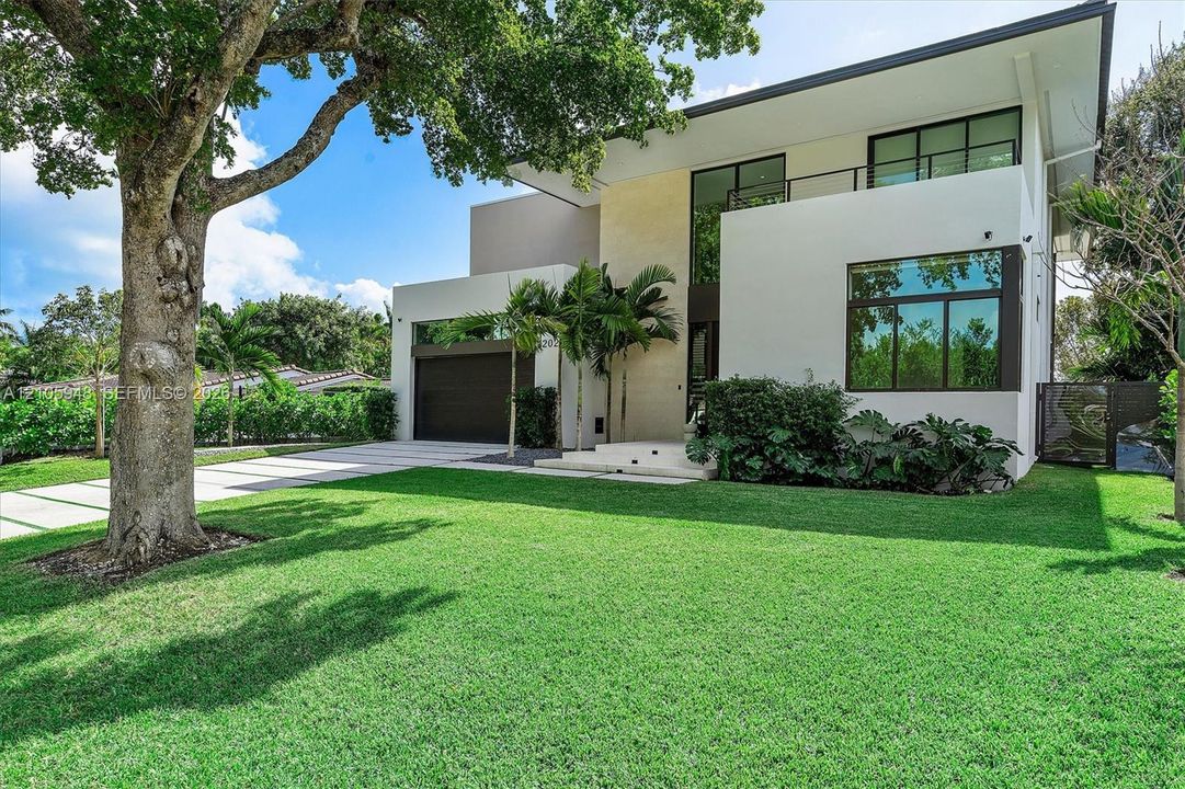 Modern two-story architectural exterior featuring smooth stucco siding, large-pane windows, a second-level balcony with metal railing, and a paved multi-car driveway