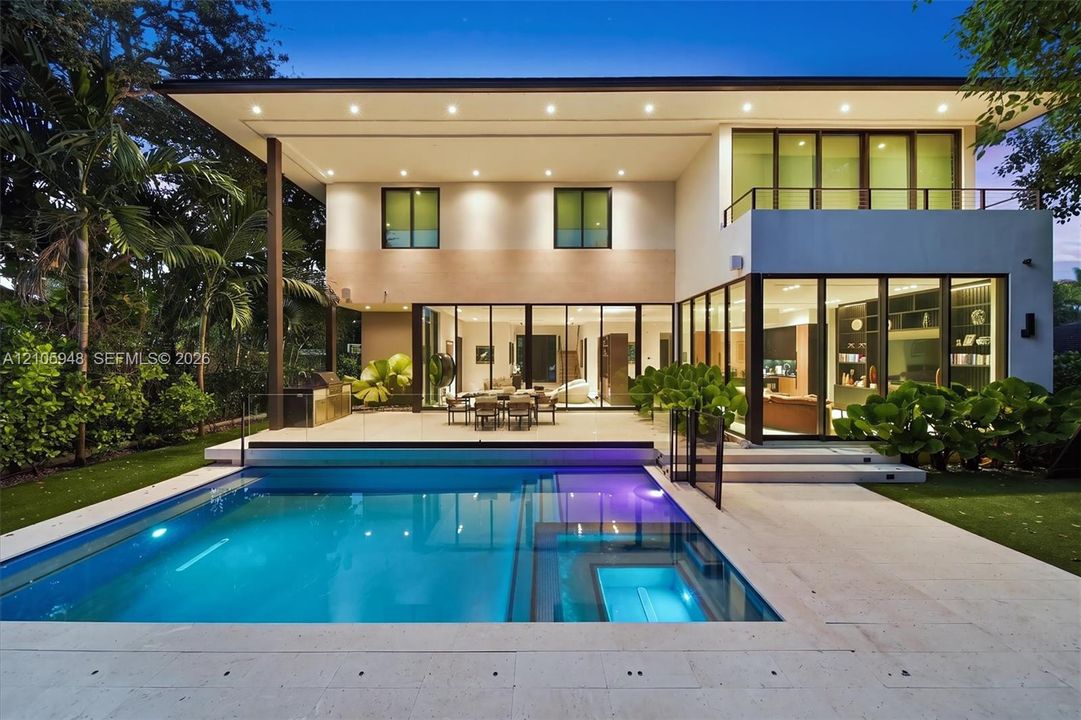 Modern two-story exterior featuring floor-to-ceiling glass sliding doors, a flat roofline, an integrated swimming pool with a stone patio surround, and a second-story balcony with a metal railing
