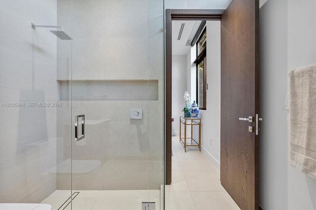 Walk-in shower featuring glass enclosure, wall-mounted rainfall showerhead, and integrated recessed shelving, complemented by a solid wood-finish door with brushed metal hardware