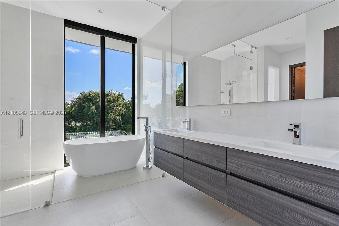Modern bathroom featuring a freestanding soaking tub, dual-sink wall-mounted vanity with dark wood-finish cabinetry, white solid-surface countertop, floor-to-ceiling glass paneling, and large-format tile flooring