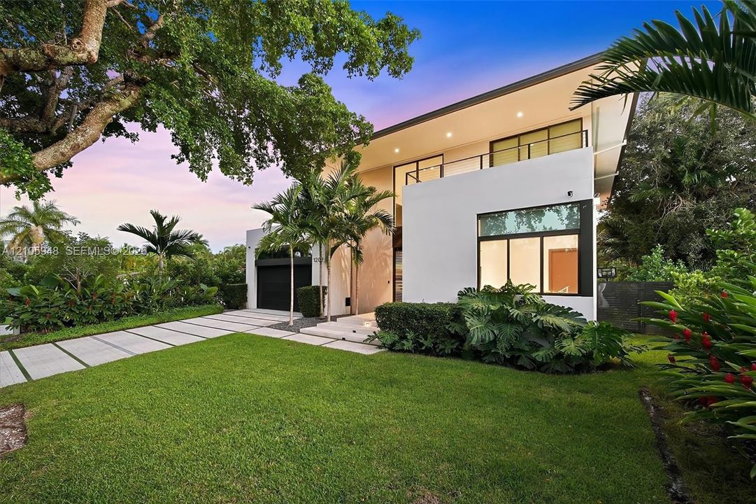 Modern architectural exterior featuring a smooth stucco facade, expansive floor-to-ceiling windows, a second-story cantilevered balcony with metal railing, and a segmented concrete paver driveway