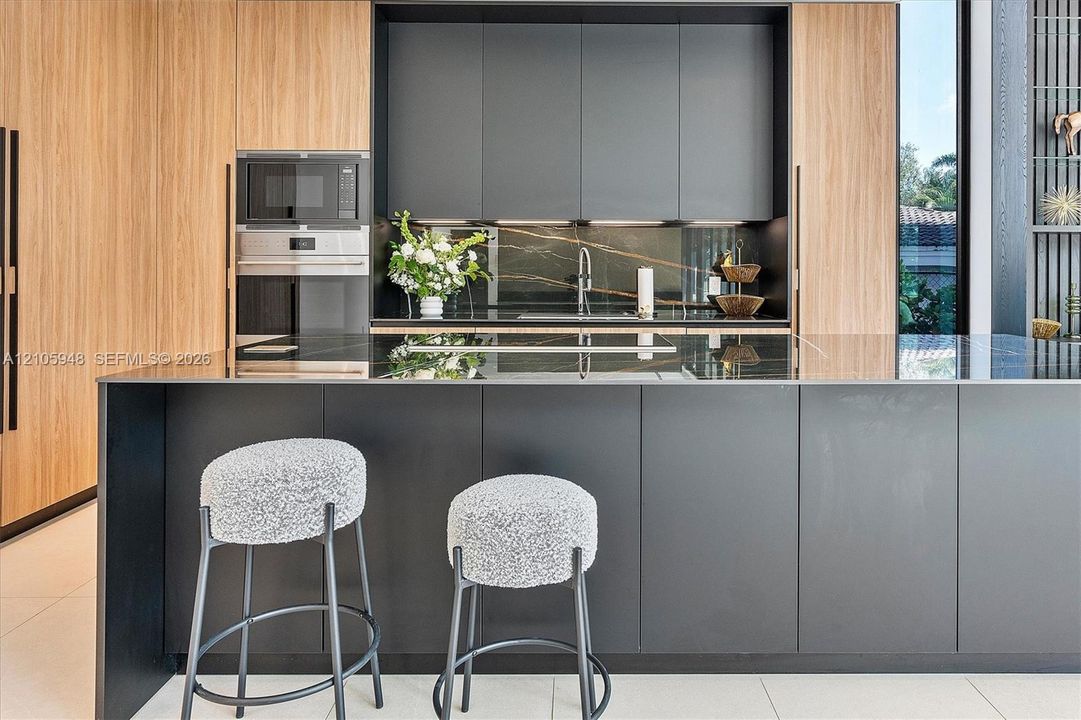 Modern kitchen featuring matte charcoal cabinetry, wood-finish paneling, an expansive island with a stone surface, built-in stainless steel appliances, and matching stone backsplash