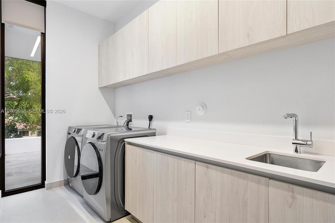 Laundry space featuring light-toned cabinetry, solid-surface countertops, an integrated stainless steel sink with a high-arch faucet, and full-height glass exterior door