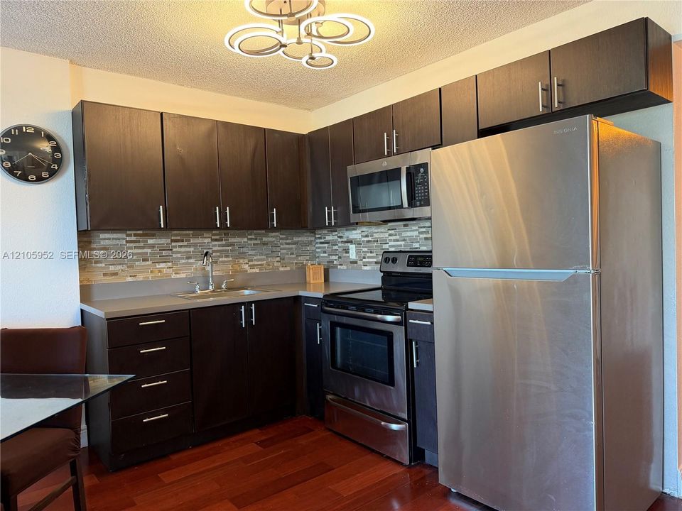 Kitchen featuring dark wood-finish cabinetry, stainless steel appliances, multi-tonal linear tile backsplash, solid-surface countertops, and a contemporary circular-ring flush-mount light fixture