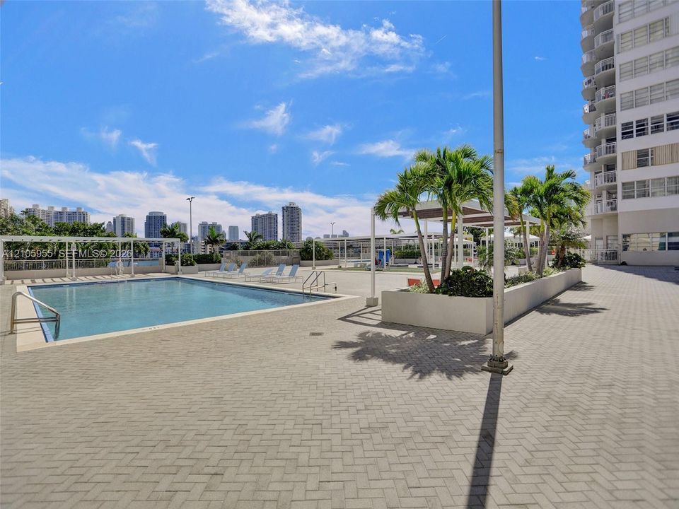 Outdoor swimming pool featuring a paved deck, built-in planter beds with mature palm trees, metal shade structures, and metal pool entry rails