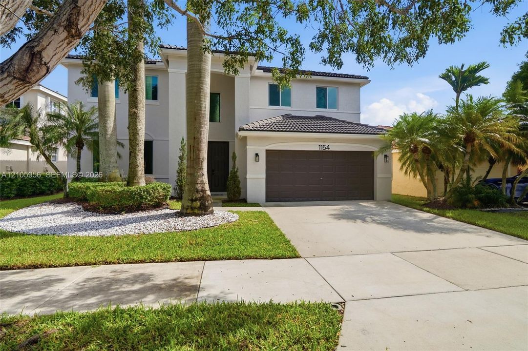 Two-story residence featuring a light-toned exterior, concrete tile roof, multi-car garage, and landscaped front grounds with mature palm trees