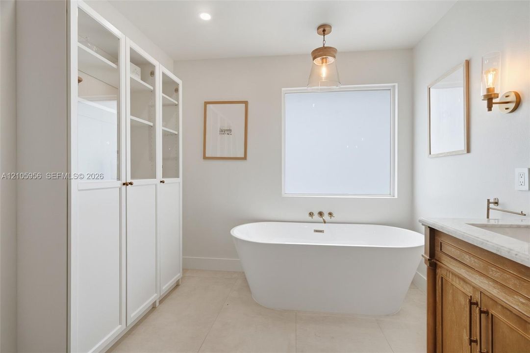 Freestanding soaking tub with wall-mounted fixtures, built-in floor-to-ceiling cabinetry with glass-panel doors, neutral tile flooring, wood-finish vanity with stone countertop, and pendant lighting