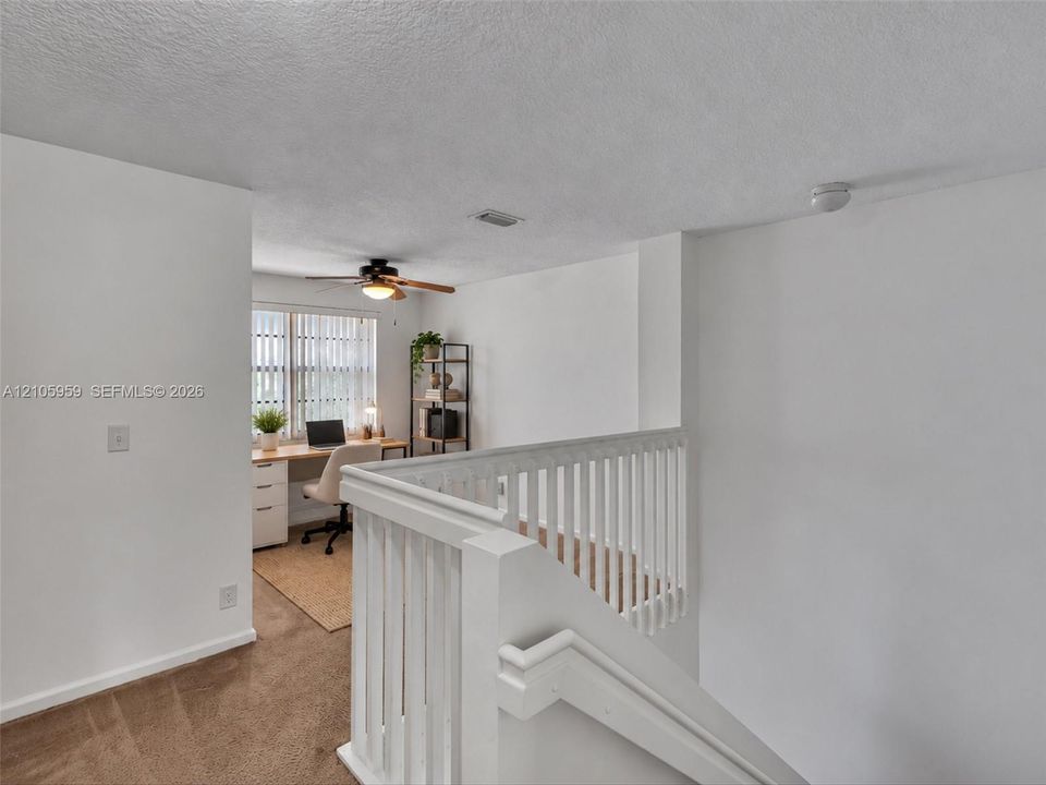 Neutral interior space featuring white walls, carpeted flooring, a white wooden staircase railing, and a ceiling-mounted fan with integrated lighting