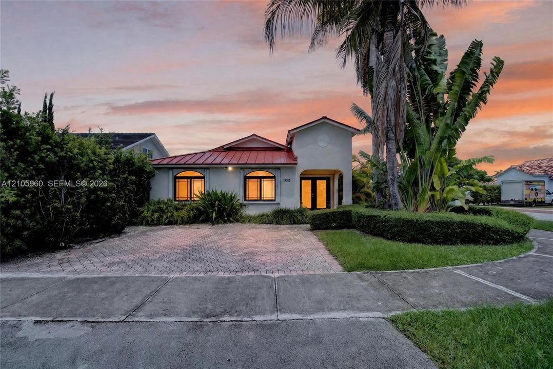 Stucco exterior featuring a red metal roof, arched windows, and a paved stone driveway, complemented by mature palm trees and manicured shrubbery