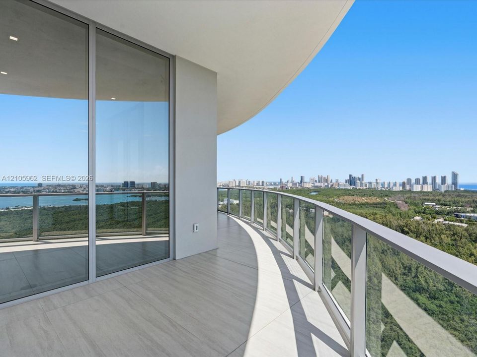 Wraparound balcony featuring light-toned tile flooring, floor-to-ceiling glass sliding doors, and a glass-paneled railing overlooking the coastline and city skyline