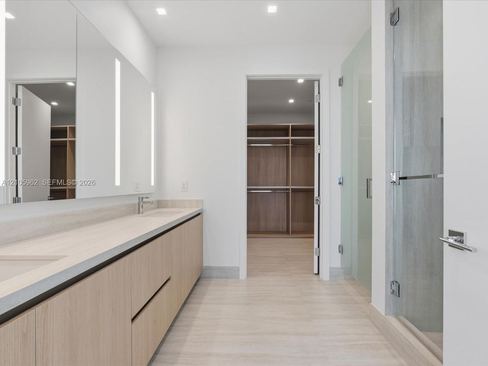 Bathroom featuring a dual-sink vanity with light wood-finish cabinetry, a large frameless mirror with integrated vertical lighting, a glass-enclosed shower, and a walk-in closet with built-in wooden shelving