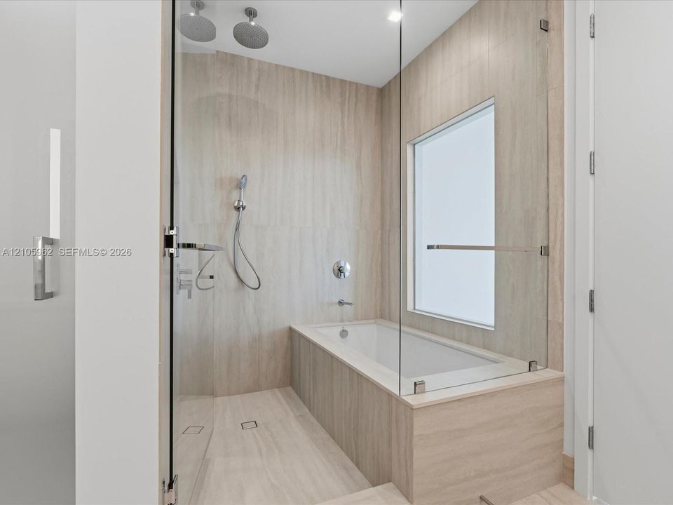 Wet room featuring large-format tile wall surrounds, a deep soaking tub, a frameless glass enclosure, dual overhead rainfall shower heads, and a wall-mounted handheld shower fixture