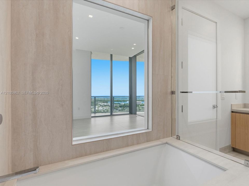 Recessed soaking tub featuring large-format stone-tile surround, clear glass enclosure, and modern metal hardware, with reflective interior glass paneling