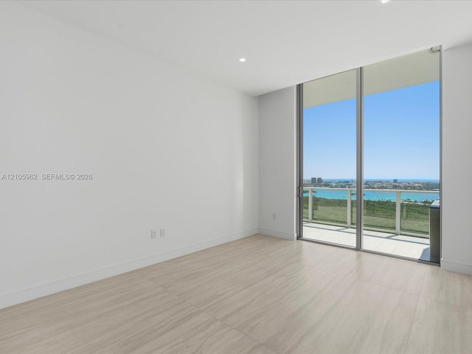 Floor-to-ceiling sliding glass doors, neutral wood-finish flooring, recessed lighting, and a glass-paneled balcony railing with water and cityscape scenery