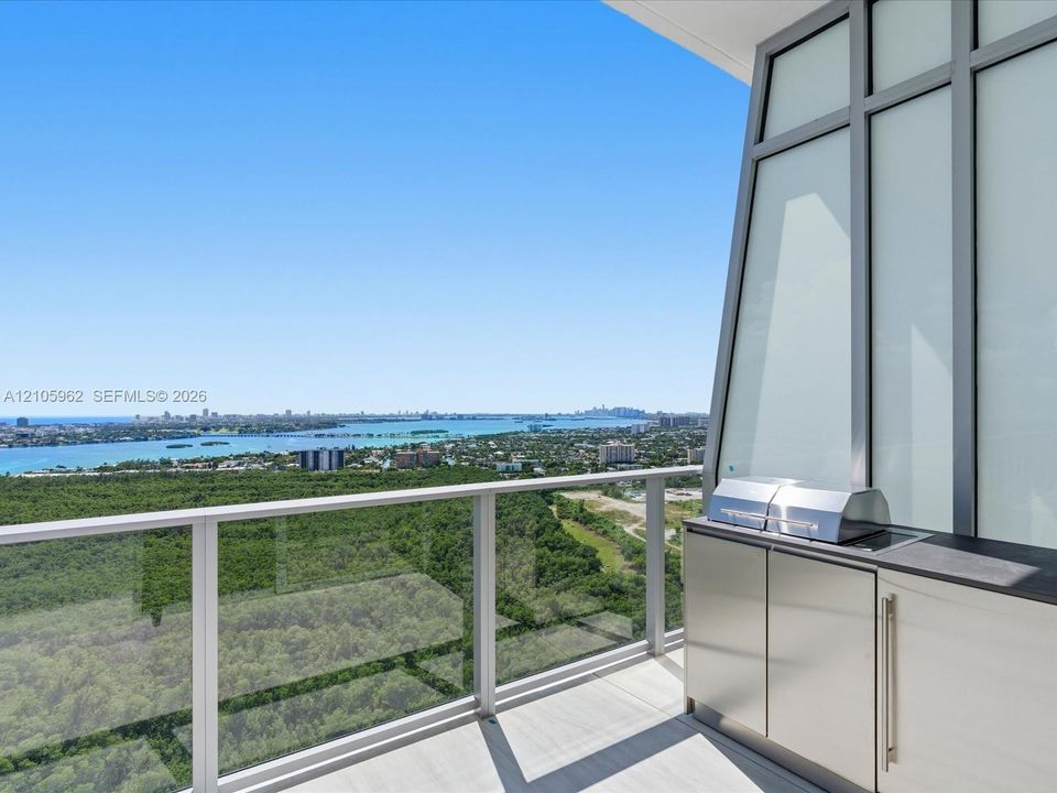 Covered terrace featuring glass panel railings, built-in outdoor kitchen with stainless steel grill, cabinetry with metal pulls, and stone-finish countertop