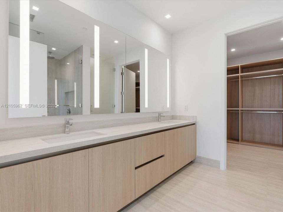 Floating double vanity with integrated sinks, neutral solid-surface countertop, floor-to-ceiling mirror with integrated LED lighting, and wood-finish cabinetry
