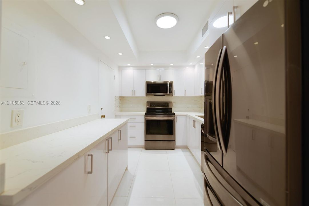 Galley kitchen featuring white cabinetry, light-toned stone-finish countertops, stainless steel appliances, recessed ceiling lighting, and large-format tile flooring