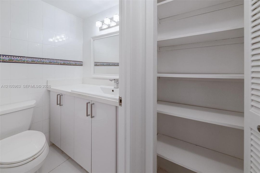 Bathroom featuring white cabinetry, integrated sink, wall-mounted mirror, ceramic tile walls with decorative border, and built-in shelving with louvered doors