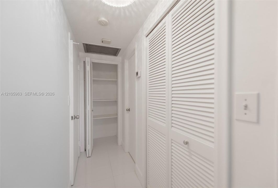 Hallway featuring white tile flooring, ceiling-mounted light fixture, and multiple sets of white louvered doors providing built-in storage