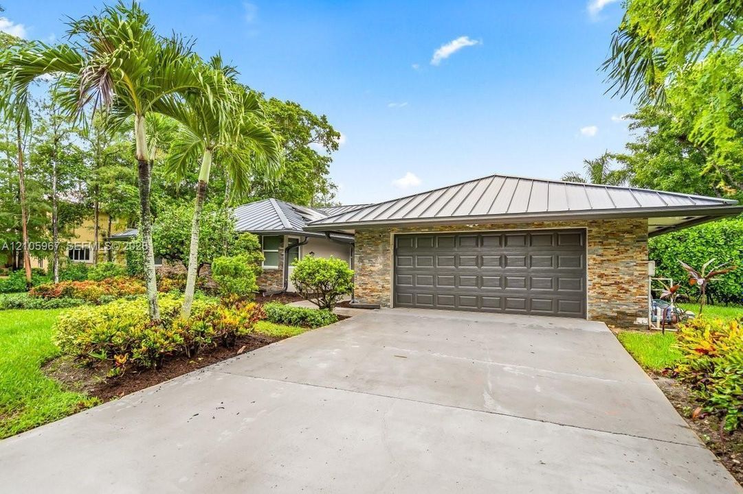 Single-story residence featuring a standing seam metal roof, stone veneer exterior, two-car garage, and concrete driveway, accented by established tropical landscaping