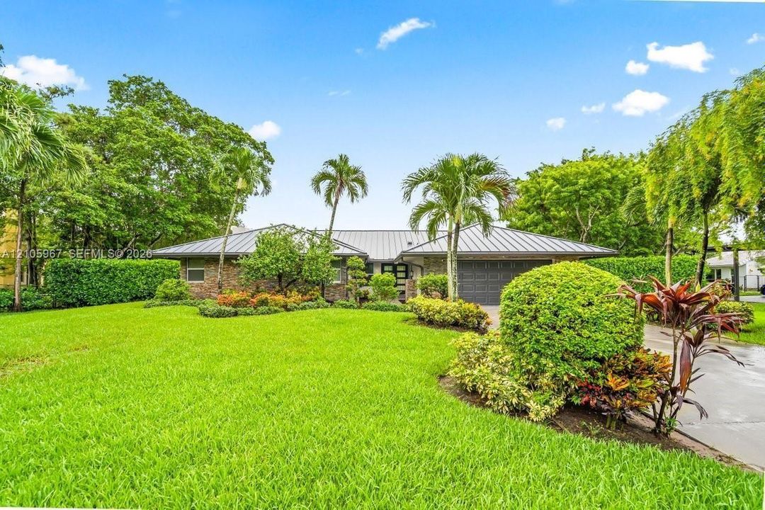 Single-story residence featuring a metal roof, brick facade, multi-car garage, and established landscaping with mature palm trees