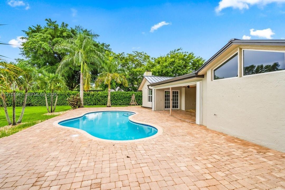 Curved in-ground swimming pool featuring a sprawling paver patio, covered porch, and exterior accent windows