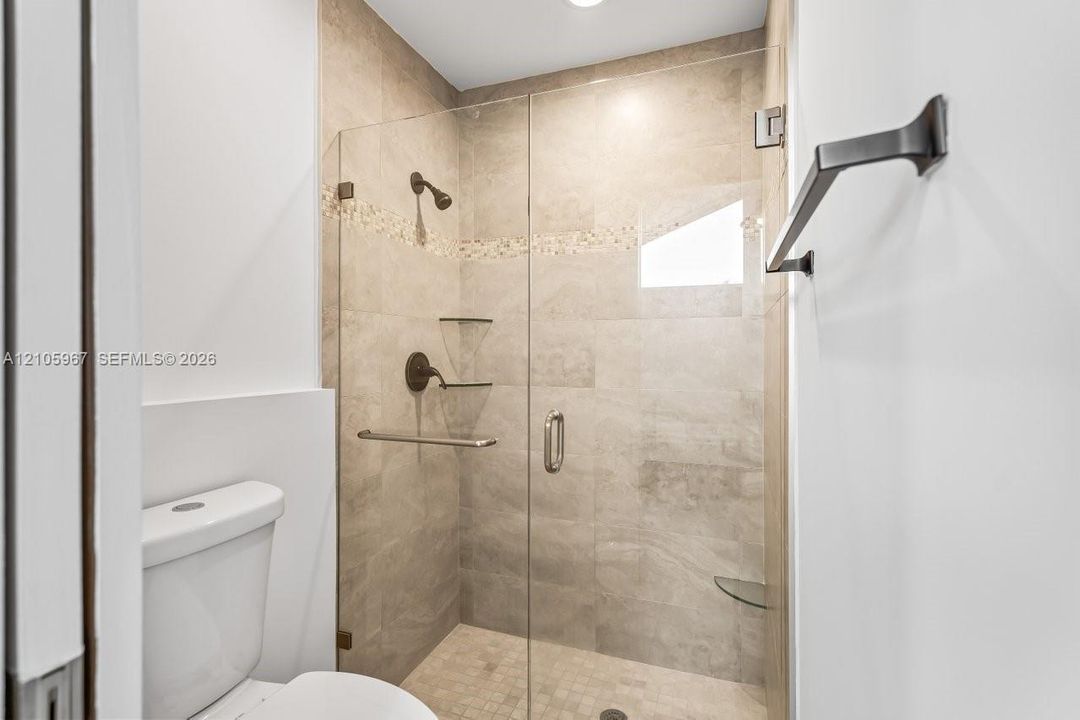 Walk-in shower featuring glass door enclosure, neutral-toned ceramic wall tile, mosaic tile accent inlay, built-in glass shelving, and matte black hardware