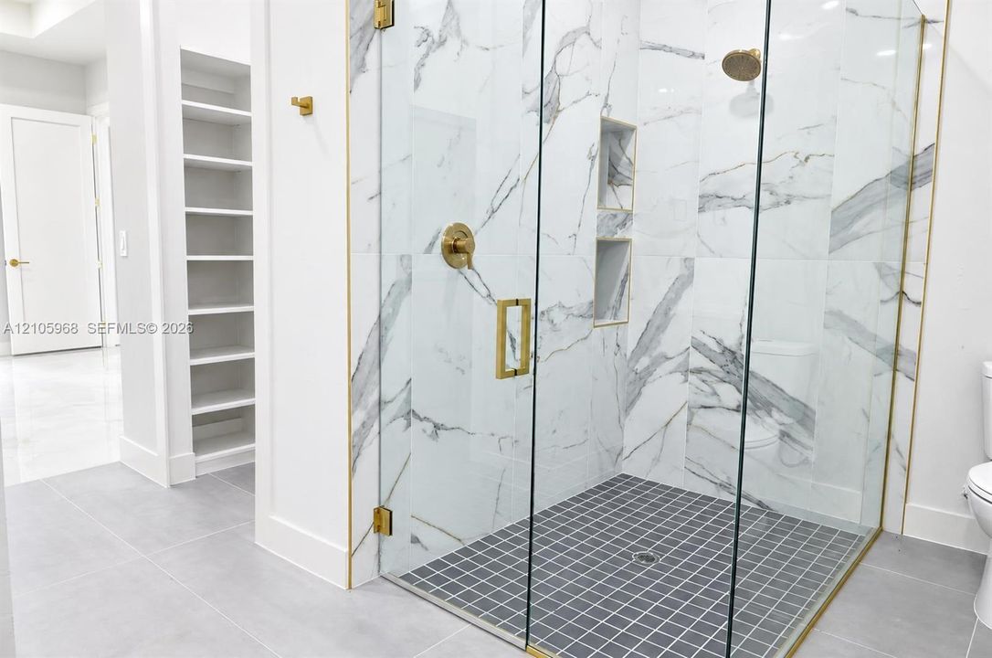 Walk-in shower featuring gold-toned hardware, floor-to-ceiling tiled walls with marbled pattern, recessed wall shelving, and a glass enclosure, complemented by built-in wall cabinetry