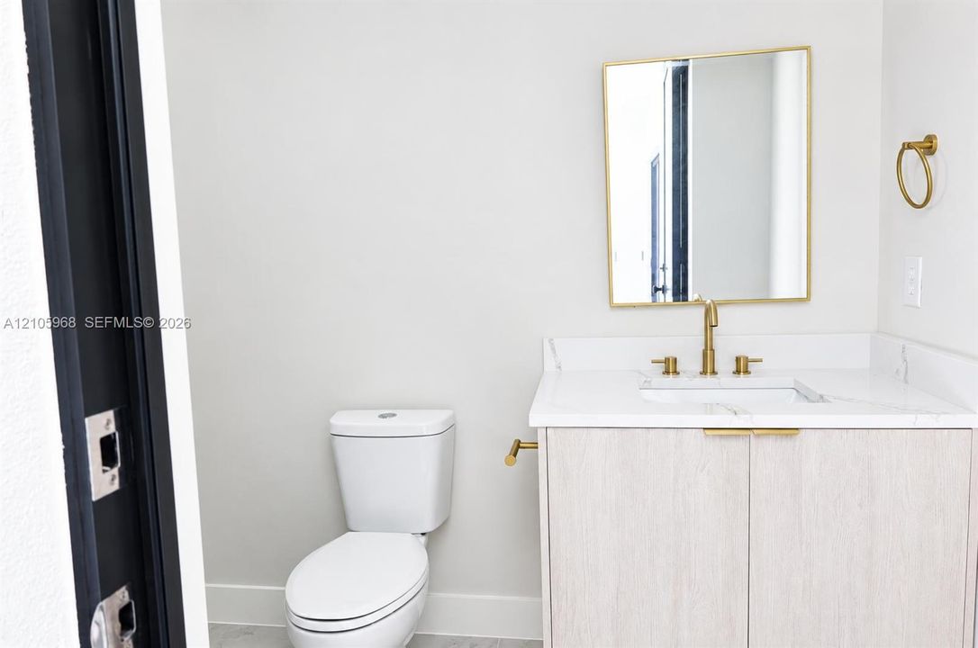 Vanity featuring a light wood-finish cabinet, stone-surface countertop, and gold-toned hardware, accompanied by a rectangular mirror with a gold-toned frame and a gold-toned widespread faucet