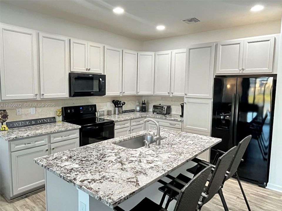 Kitchen featuring light-toned cabinetry, patterned stone-finish countertops, central island with an undermount sink, recessed lighting, and black appliances