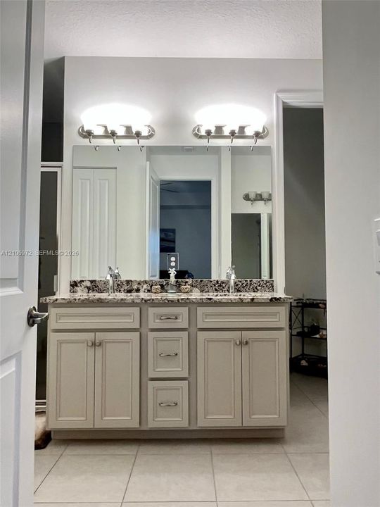 Double-sink vanity featuring neutral-toned cabinetry, a stone-patterned countertop, a large wall-mounted mirror, and dual light fixtures