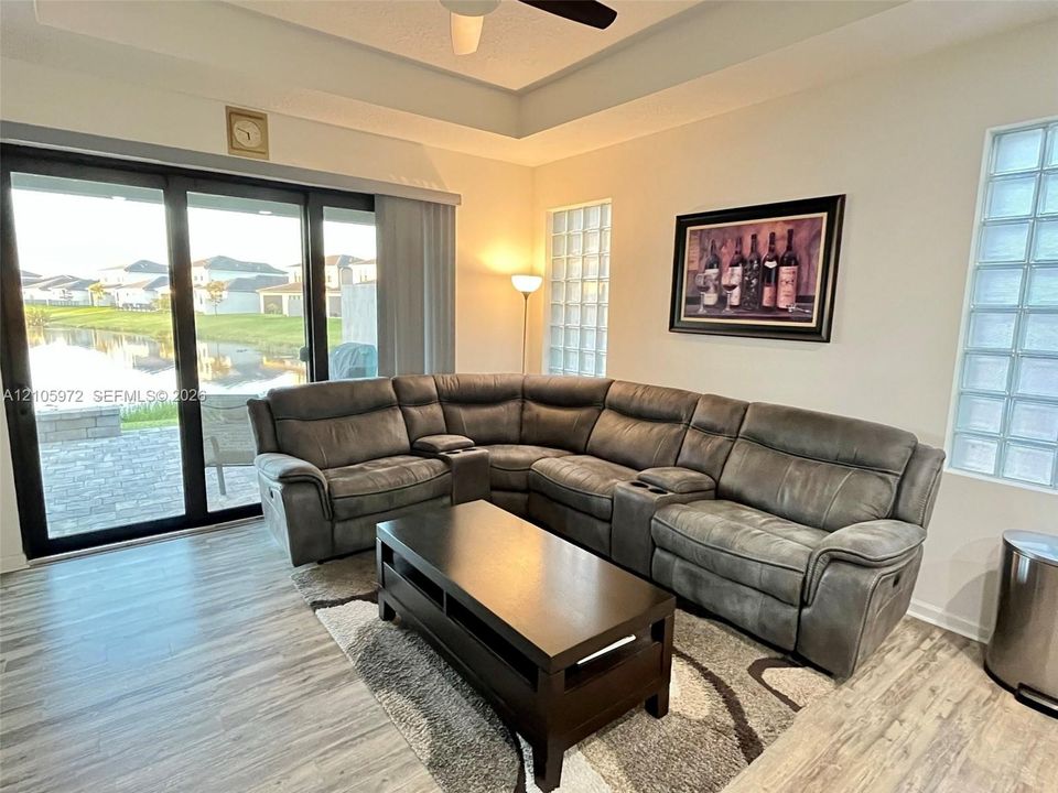Tray ceiling with integrated fan, wood-finish flooring, and glass block windows, featuring large sliding glass doors opening to a paved patio area