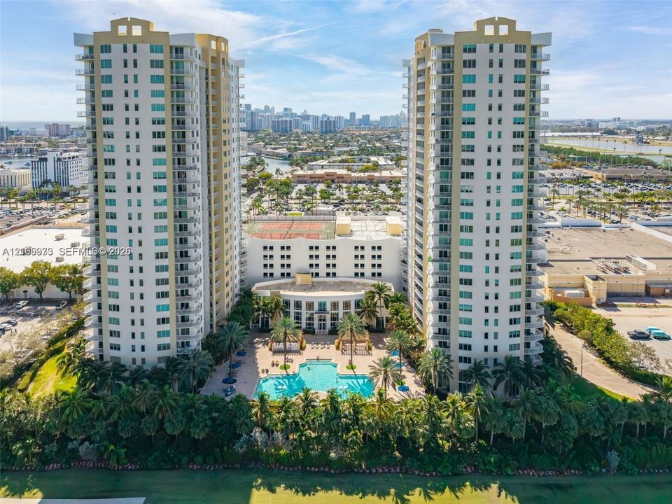High-rise residential towers featuring multiple wrap-around balconies, a ground-level swimming pool, hardscaped patio areas, and tennis courts