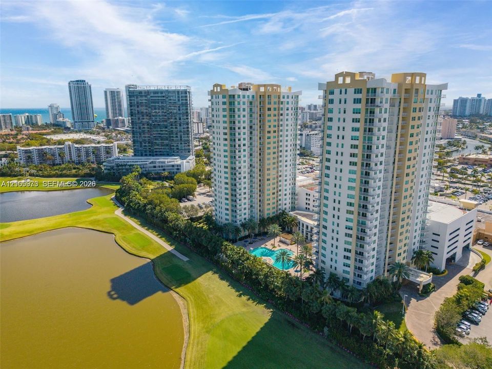 High-rise residential towers featuring balconies, ground-level swimming pool, and dedicated parking structure, positioned along a water feature and golf course
