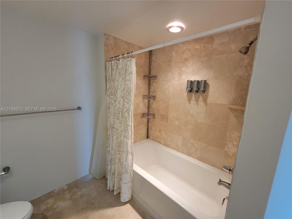 Bathtub with full-height stone tile surround, integrated metal shelving, wall-mounted shower head, and recessed lighting