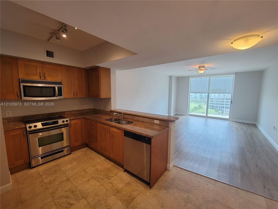 Kitchen featuring wood cabinetry, stone countertops, stainless steel appliances, and tile flooring, opening to a space with wood-finish flooring and floor-to-ceiling windows