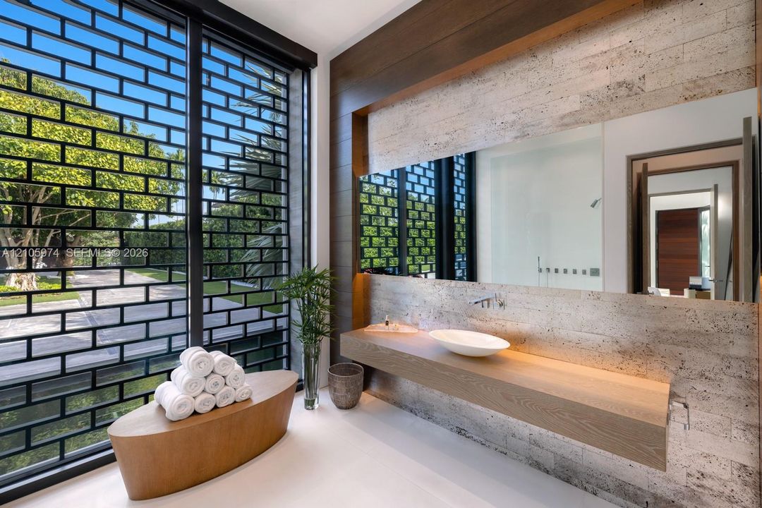 Floating wood-finish vanity with a white vessel sink, wall-mounted faucet, oversized frameless mirror, textured stone-tiled wall, and full-height exterior window with integrated metal grid screen