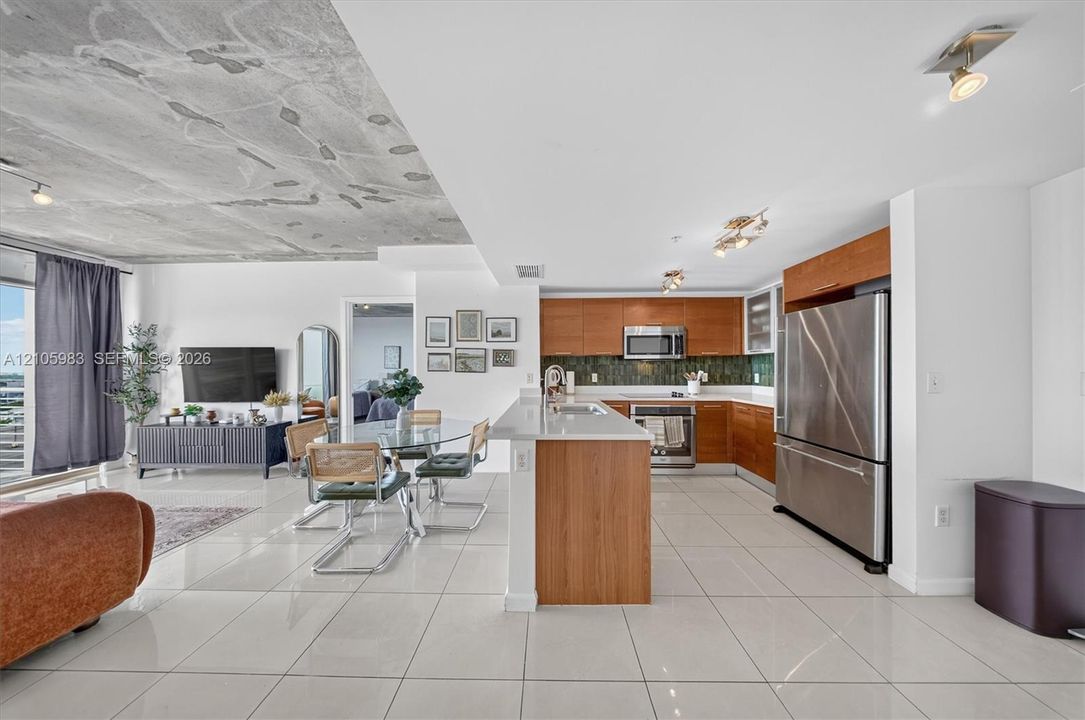 Open-concept kitchen featuring wood-finish cabinetry, stainless steel appliances, quartz countertops, a decorative tile backsplash, and large-format porcelain tile flooring throughout