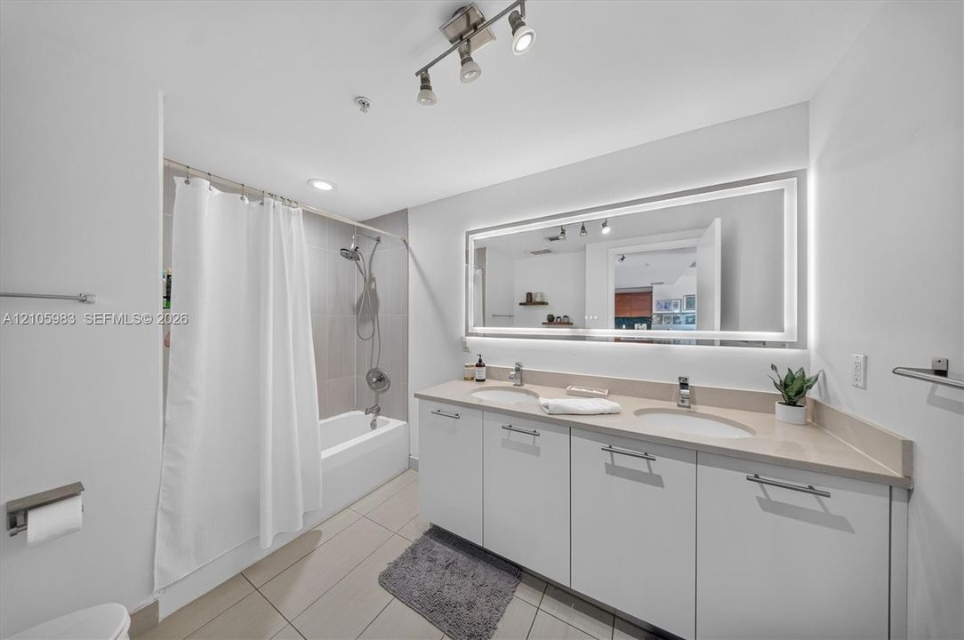 Double vanity featuring integrated sinks, light-toned solid surface countertops, flat-panel cabinetry, a large illuminated mirror, and a tub-shower combination with tile surround