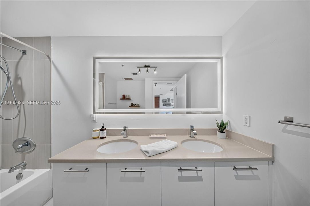 Dual-sink vanity with a solid-surface countertop, white cabinetry with horizontal hardware, backlit rectangular mirror, and a tile-surround shower and tub combination