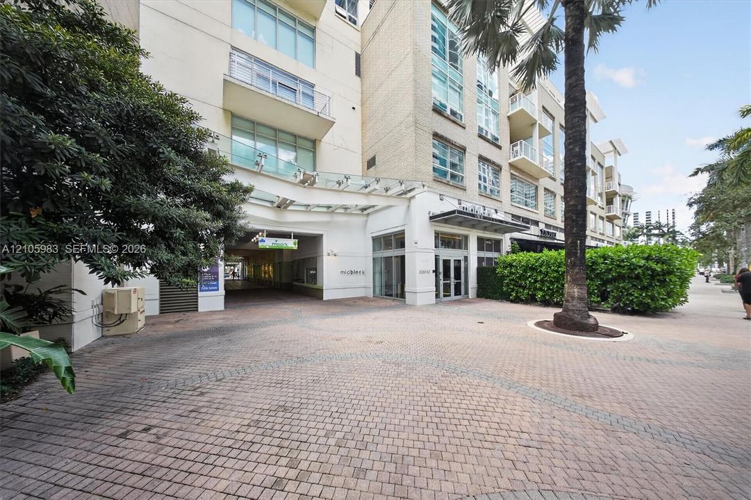 Exterior building facade featuring a glass and steel porte-cochère, multi-level balconies, masonry construction, and a paved brick driveway